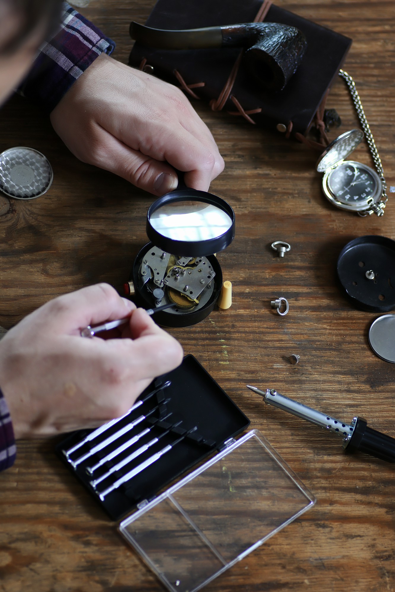 Mani al banco che osservano con una lente il movimento di un orologio da tasca, tra cacciaviti di precisione